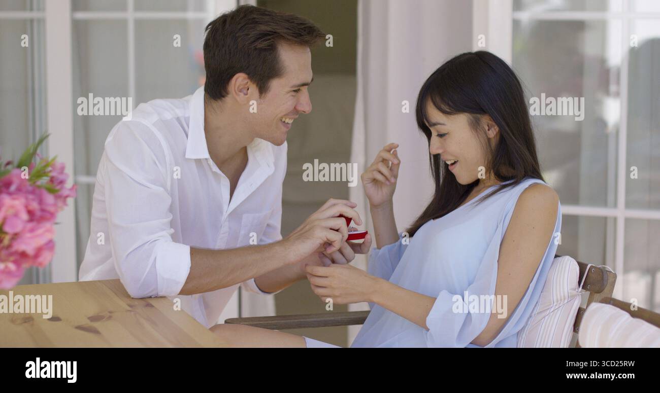 Giovane eccitato con camicia bianca che dona alla donna lusingata una camicetta blu un anello all'interno della cassa a tavola Foto Stock