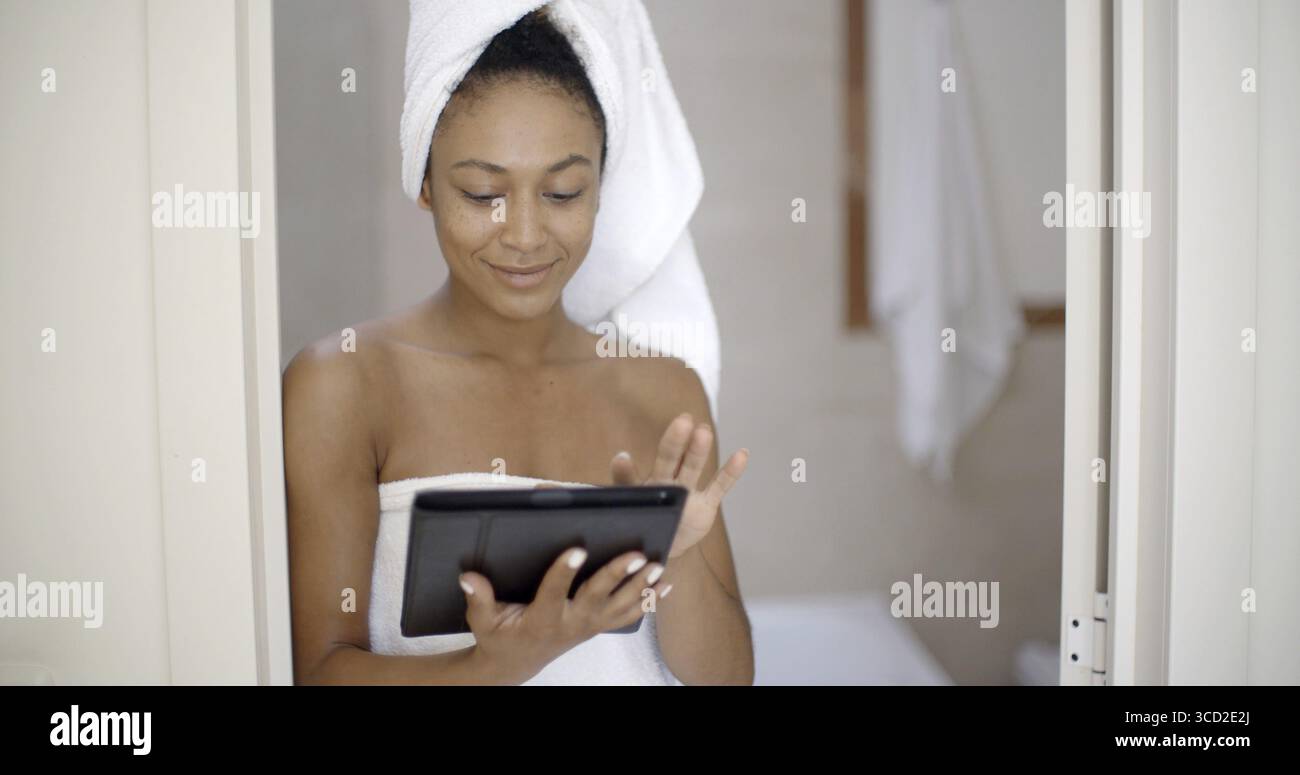 Attraente giovane donna nera con un tablet touchscreen appoggiato sulla porta del bagno Foto Stock