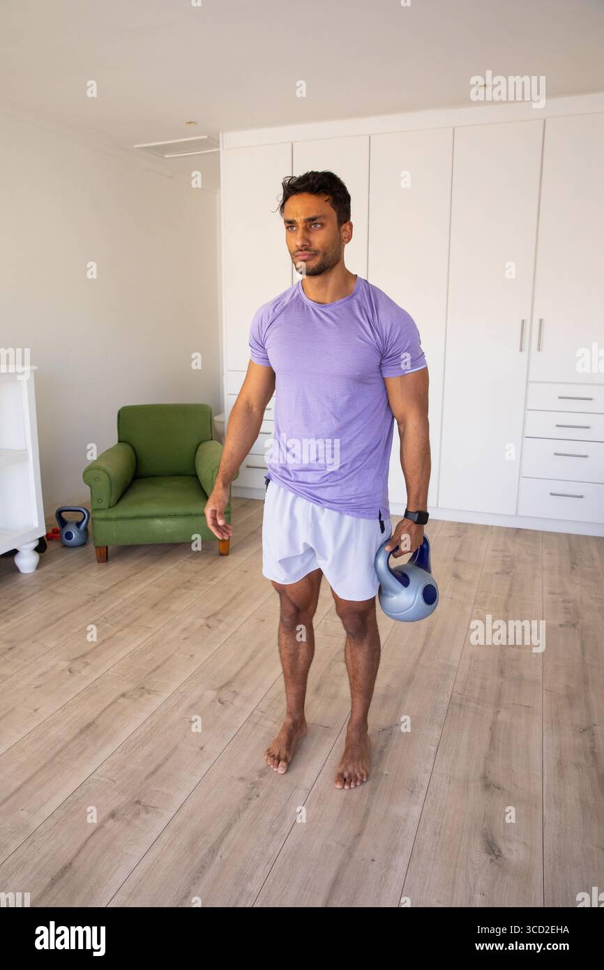 Uomo indiano con kettlebell e smartwatch in piedi in palestra vicino alla poltrona verde Foto Stock