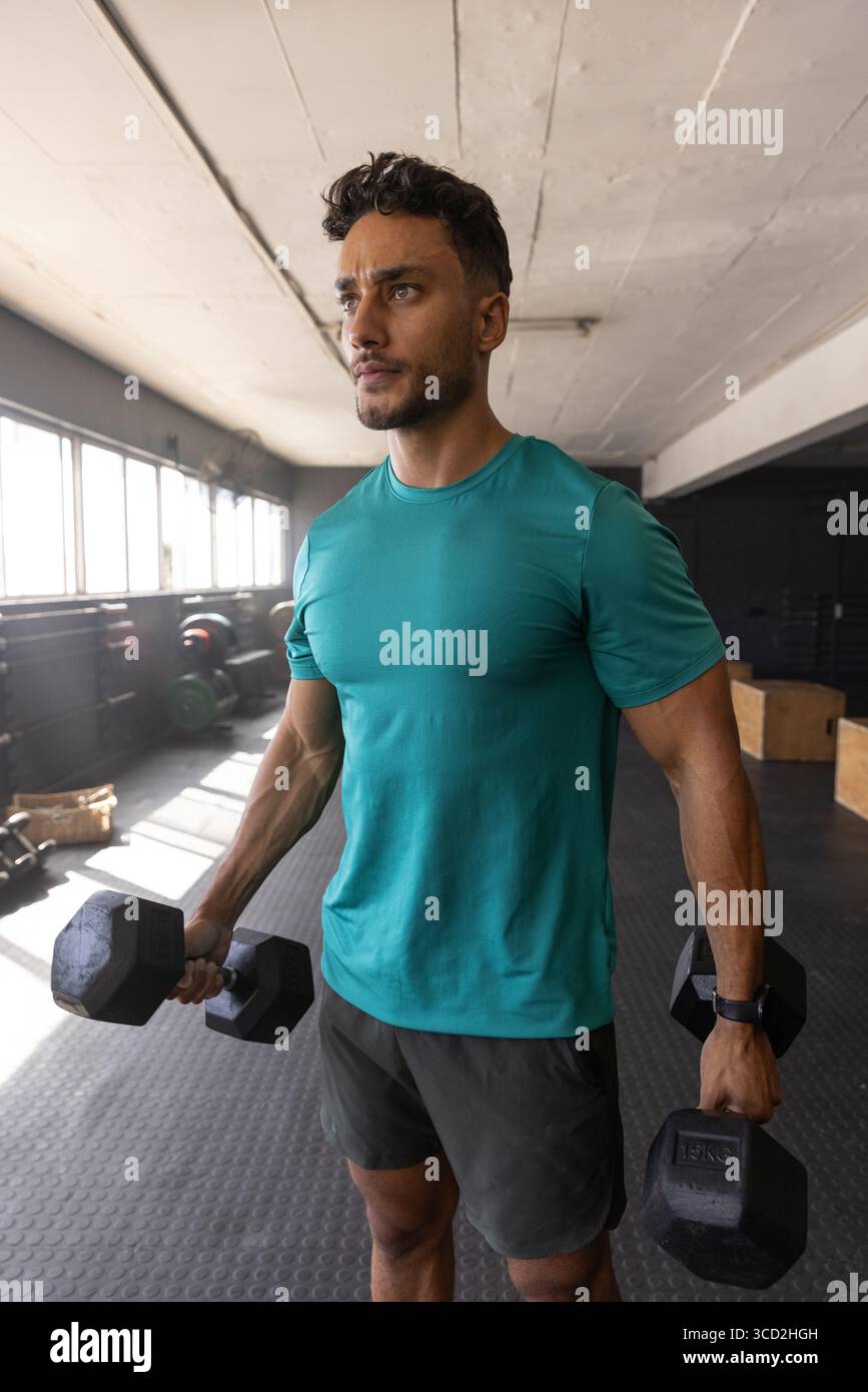 Uomo del Medio Oriente che indossa una camicia color tè, pantaloncini neri che sollevano manichini esagonali in palestra vicino alle piastre per pesi Foto Stock
