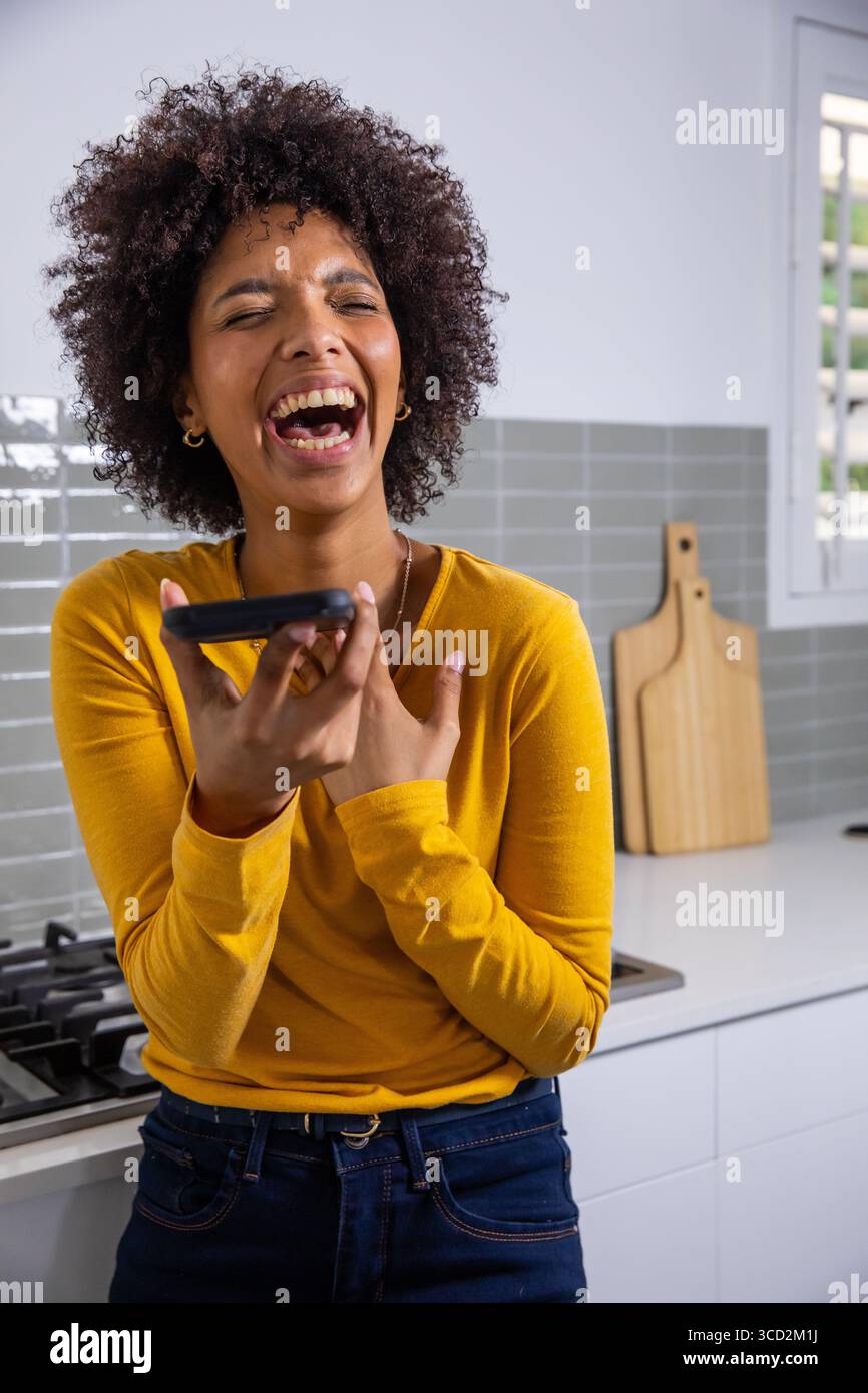 Donna afroamericana che ride e parla allo smartphone in cucina con taglieri Foto Stock