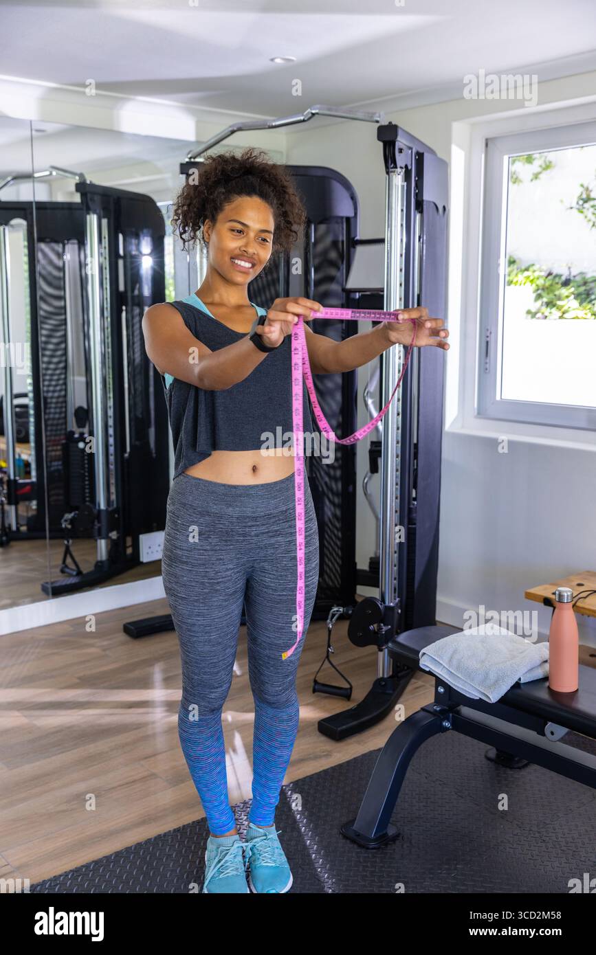 Donna afro-americana che sorride mentre tiene il metro a nastro in sala fitness con la puleggia del cavo Foto Stock