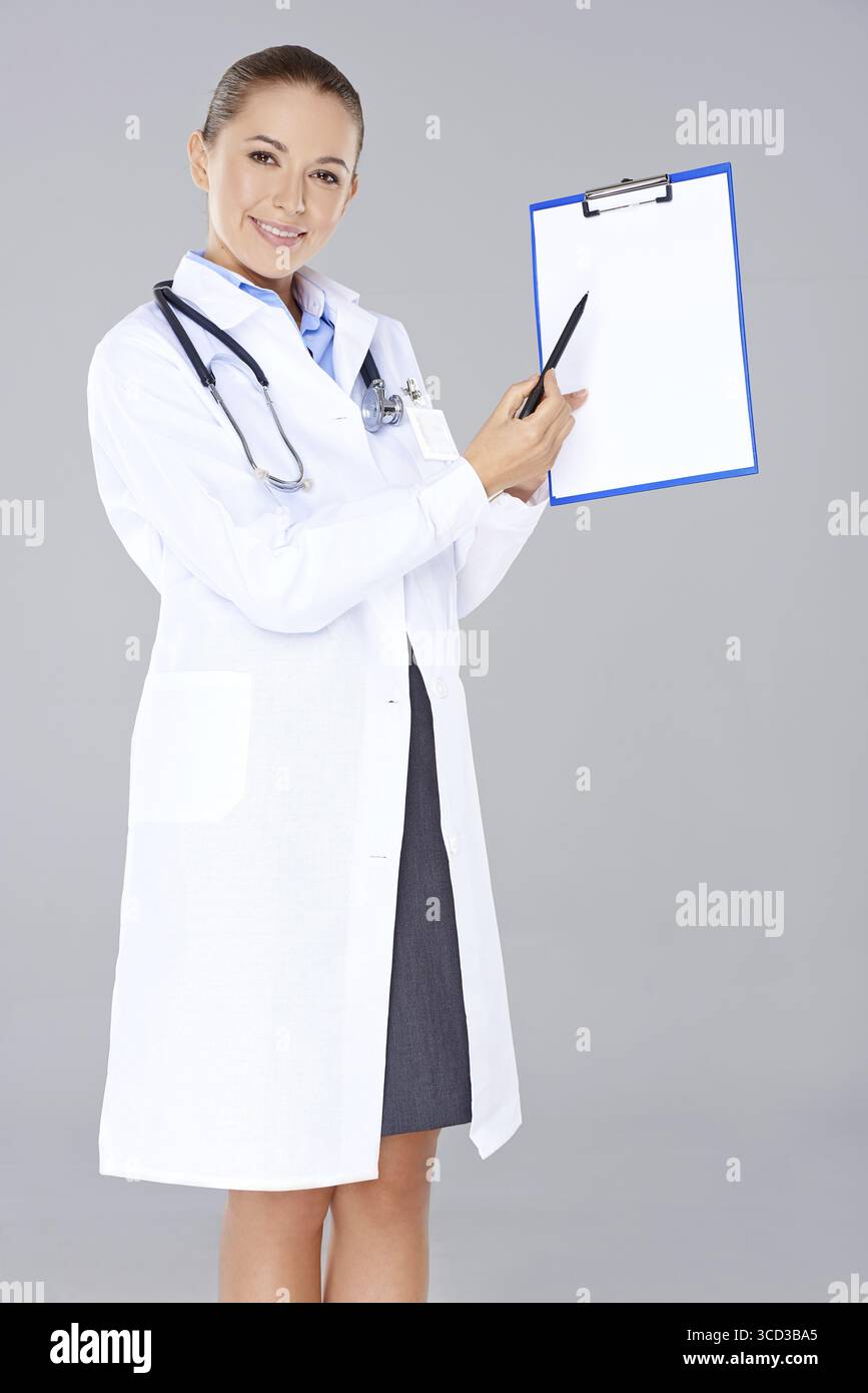 Attraente giovane medico o infermiera con un camice da laboratorio e uno stetoscopio che indica un portadocumenti vuoto che tiene in mano con un sorriso sul grigio Foto Stock