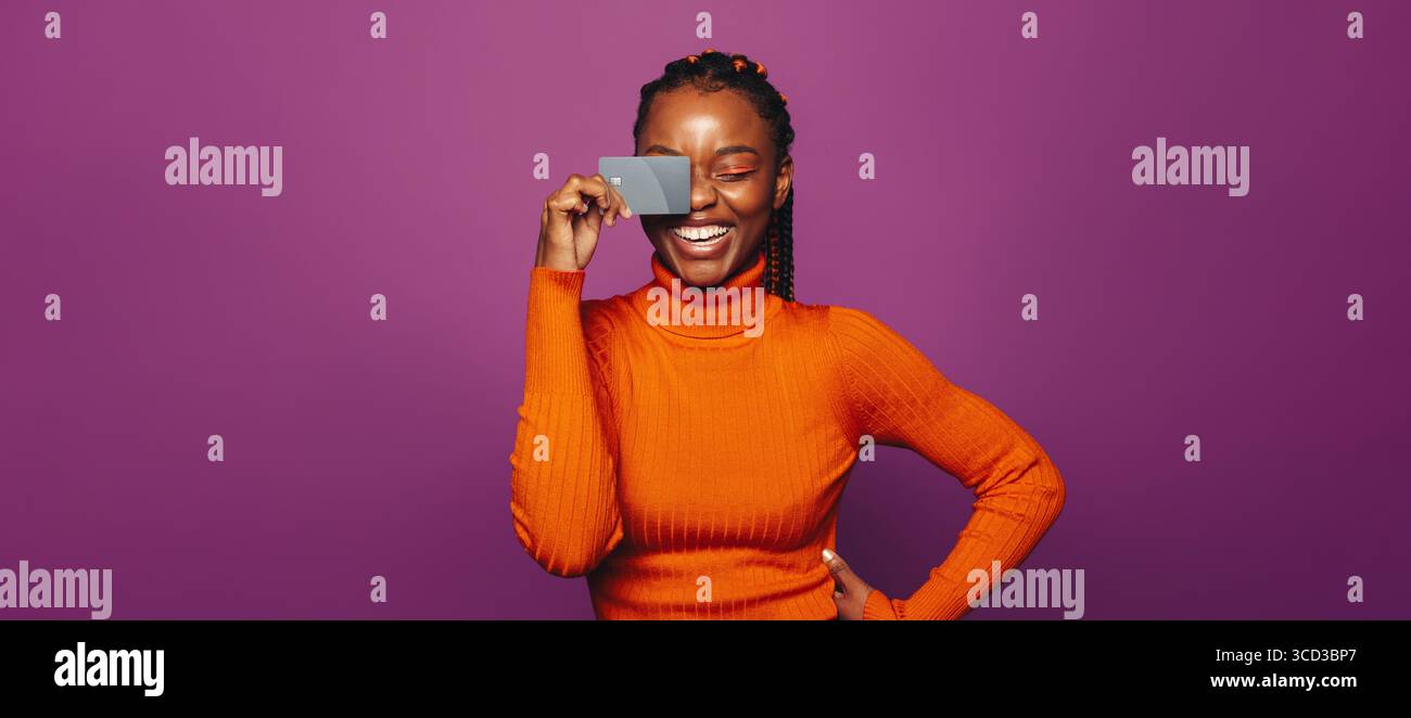 Giovane donna felice che tiene in mano una carta di credito in faccia mentre sorride in un colorato ambiente da studio. Concetto di shopping online, pagamenti digitali. Foto Stock