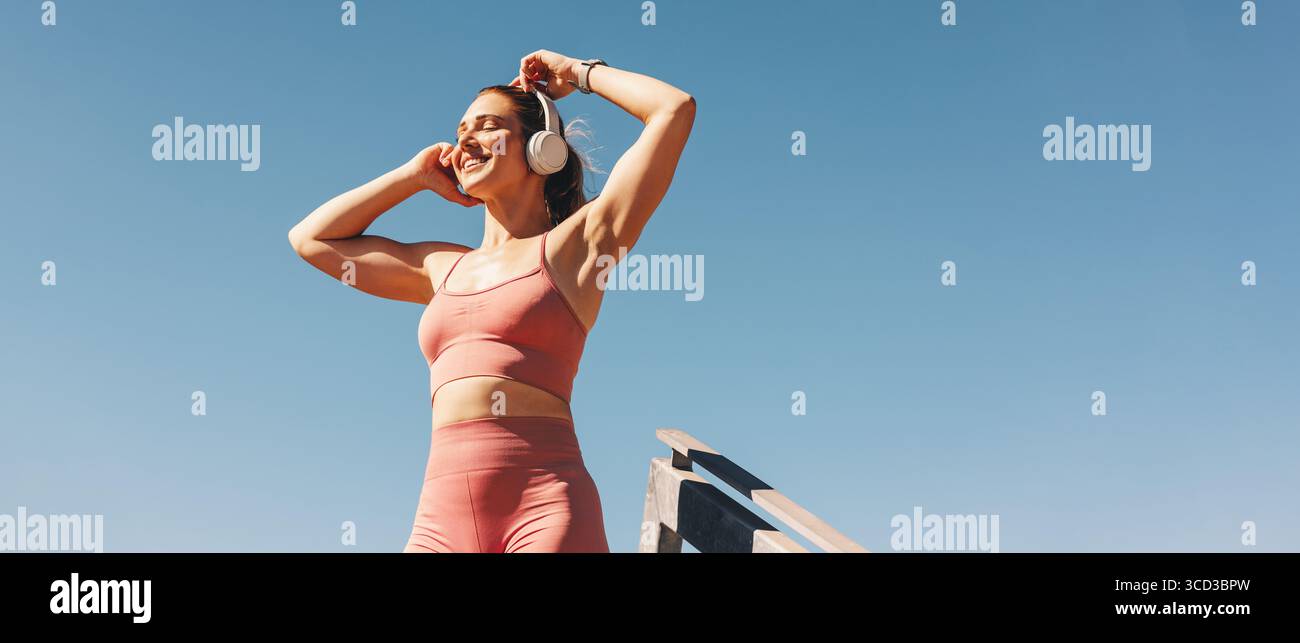 Indossate una giovane donna che indossa le cuffie e gli abiti da ginnastica, ascoltando la musica mentre si trova all'aperto sotto un cielo limpido. Salute, fitness, motivazione. Foto Stock