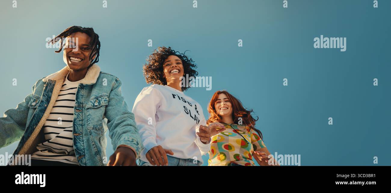 Tre giovani amici alla moda che ridono e camminano insieme sotto un cielo limpido. Sottolinea la libertà, la connessione sociale e la vibrante energia dei giovani. Foto Stock