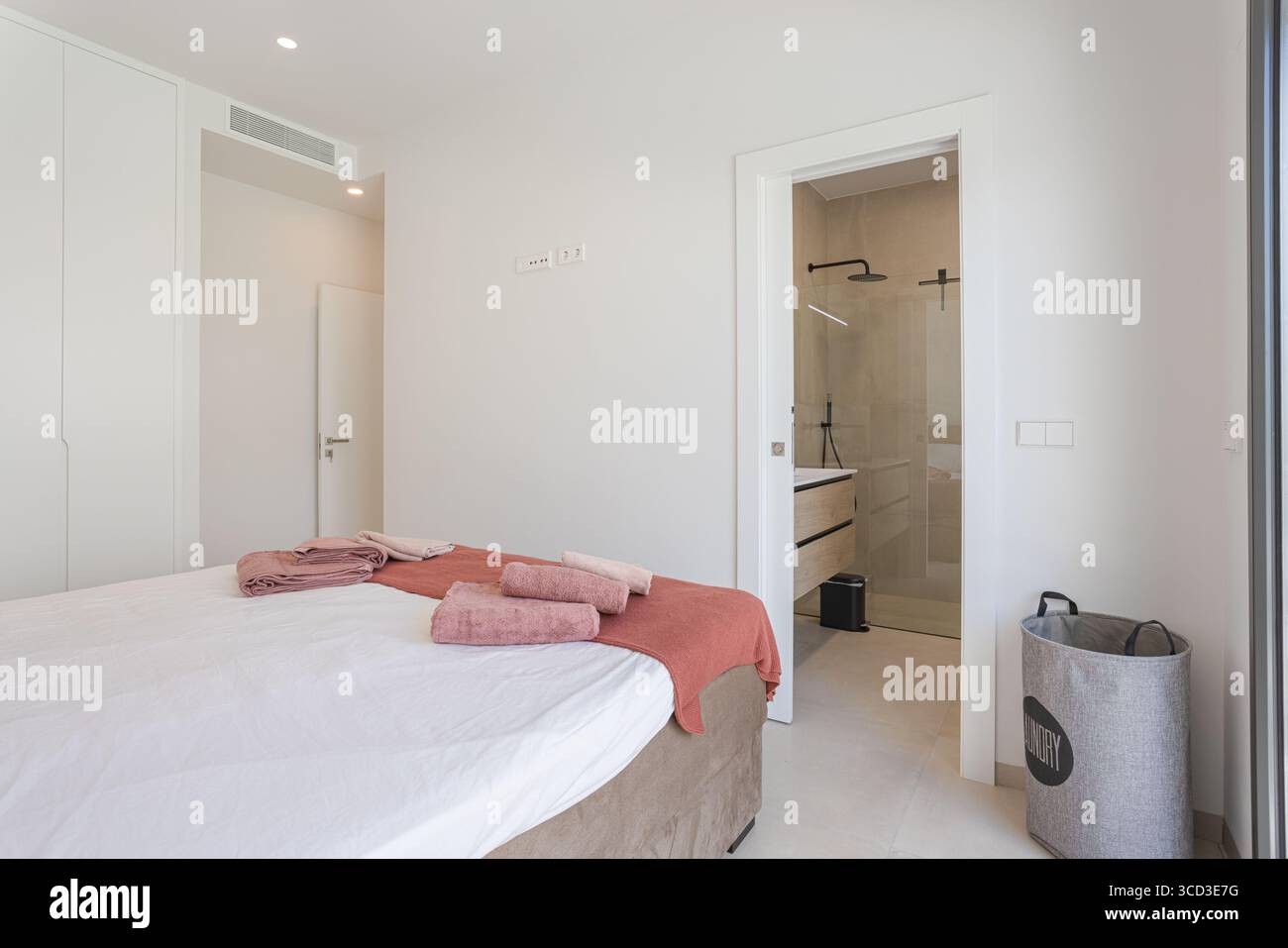 Camera luminosa con pareti bianche, morbidi asciugamani rosa e accesso diretto a un moderno bagno privato con cabina doccia. Foto Stock