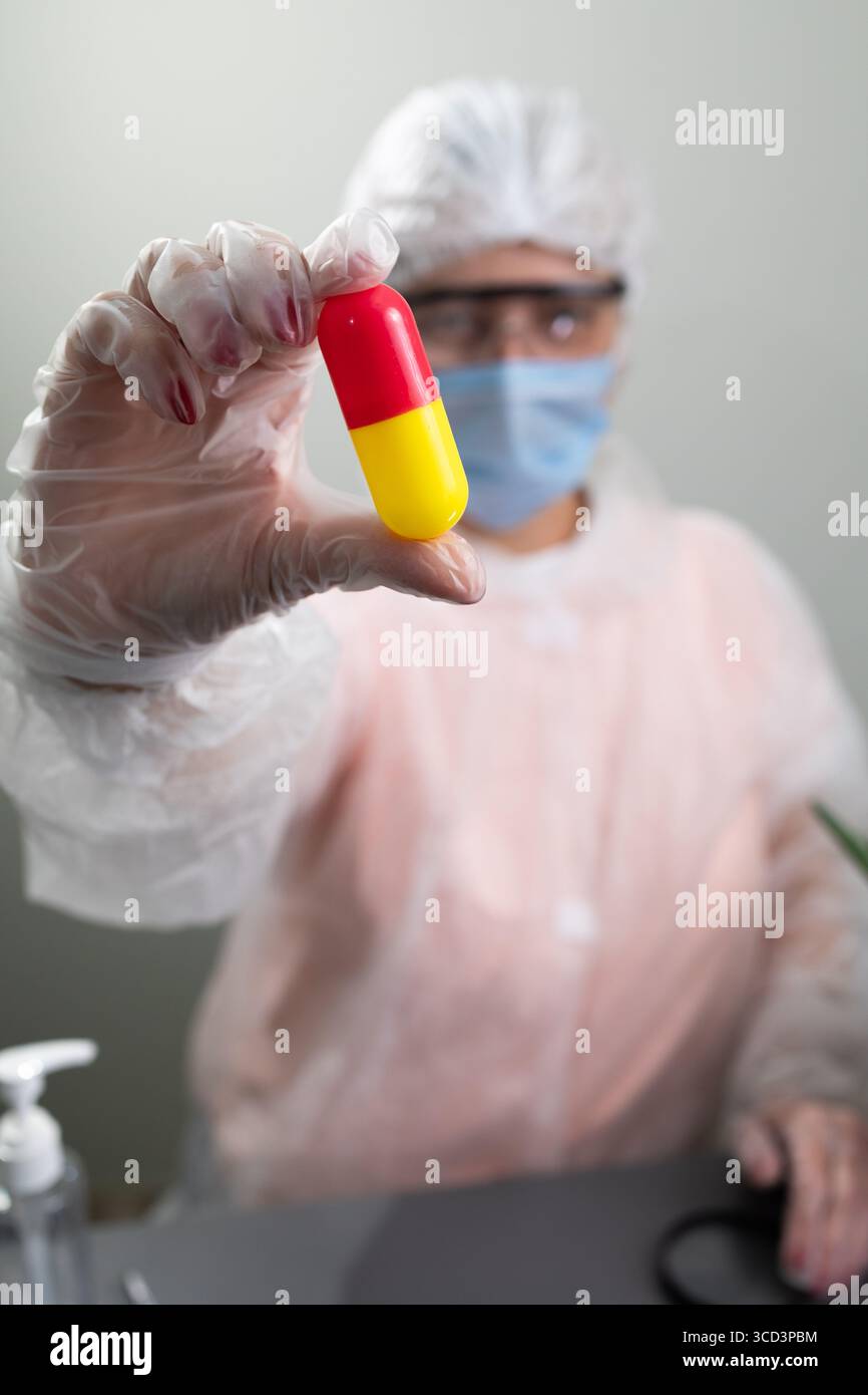 Un ricercatore indossa l'equipaggiamento protettivo mentre tiene in mano una capsula rossa e gialla, concentrandosi sullo studio delle applicazioni dell'aloe in medicina. L'ambiente suggerisce un ambiente di laboratorio. Foto Stock