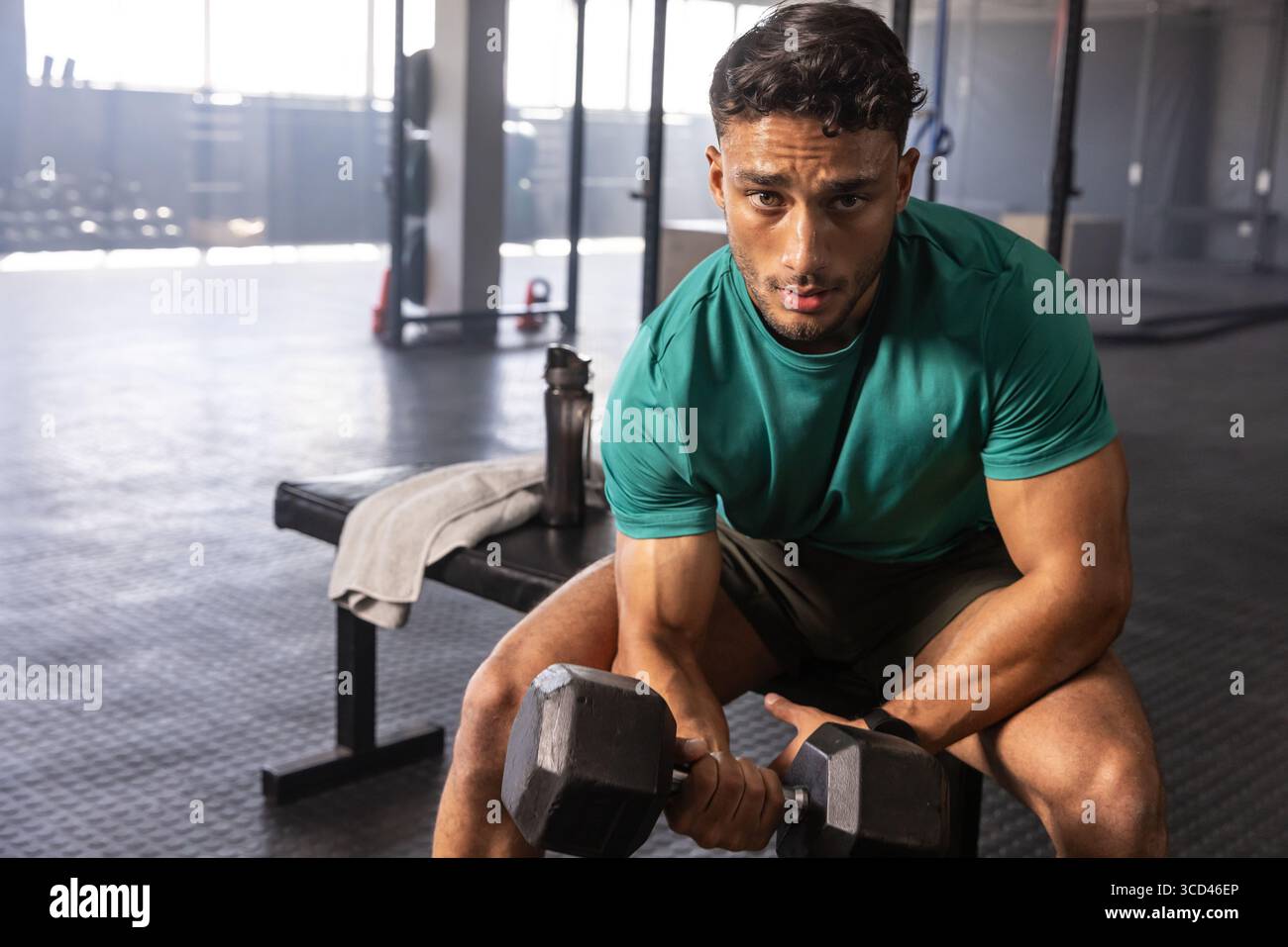 Uomo muscoloso che solleva il manubrio sulla panchina della palestra tra rack e campanelli, usando un asciugamano e una bottiglia d'acqua Foto Stock