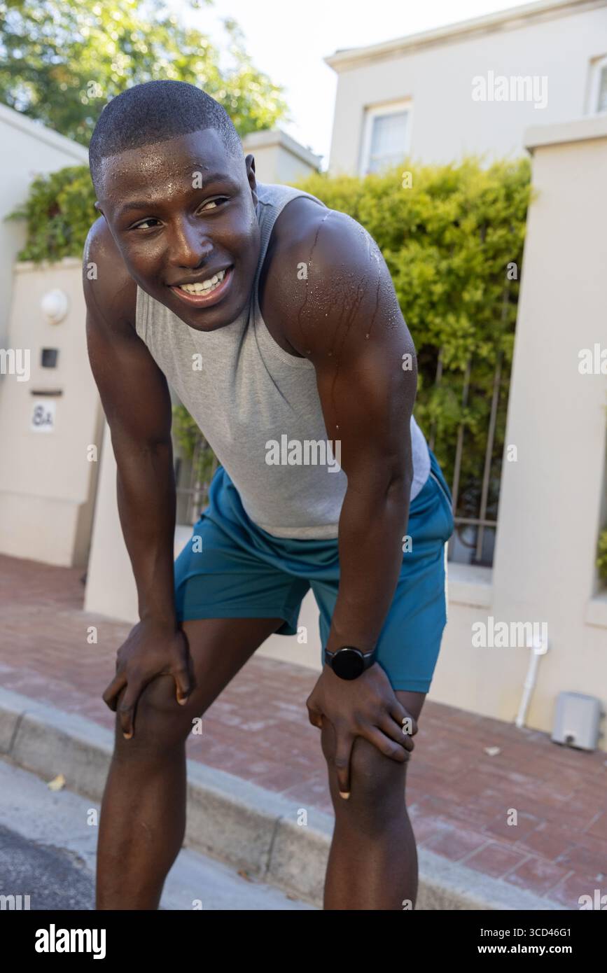 Atleta afroamericano che si appoggia sul marciapiede e indossa canotta, pantaloncini corti con orologio sportivo nero Foto Stock
