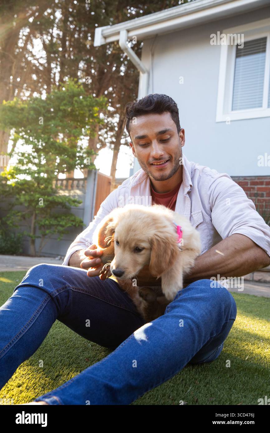 Uomo seduto su un tappeto erboso artificiale che tiene un cucciolo dorato con colletto rosa davanti al dorso grigio Foto Stock