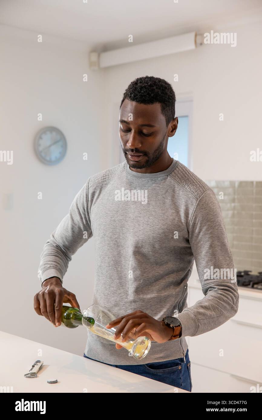 Uomo afroamericano che versa la bevanda dalla bottiglia verde al vetro in cucina accanto all'apripista, tappo Foto Stock