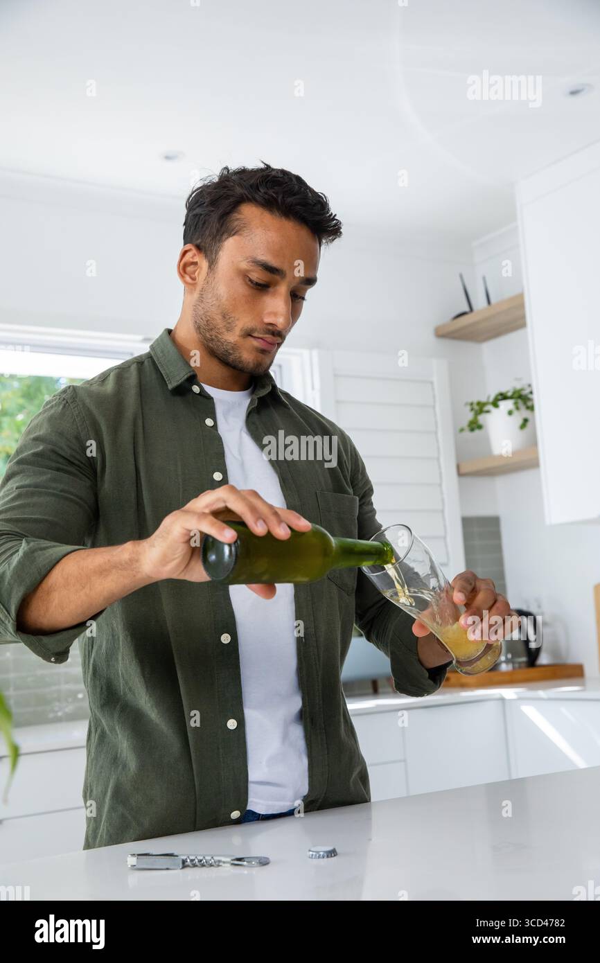 Uomo che versa la bevanda da una bottiglia verde in un alto bicchiere all'isola della cucina con apripista e tappo Foto Stock