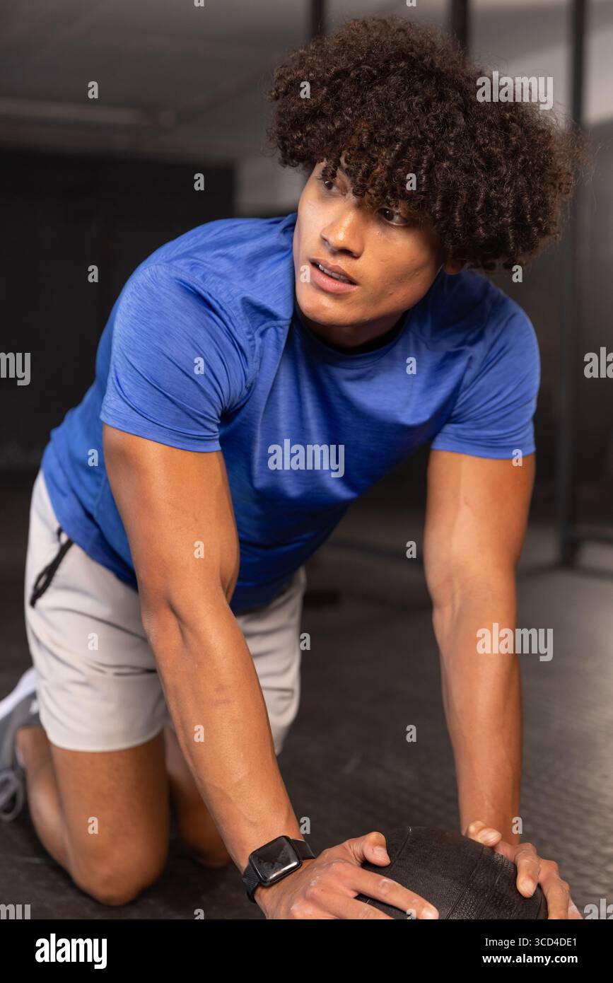 Uomo con camicia atletica blu inginocchiato sul pavimento in gomma della palestra premendo palla medica vicino allo squat rack Foto Stock