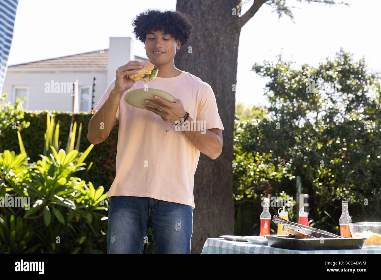 Uomo afroamericano in piedi nel cortile tenendo la piastra, sollevando l'hamburger vicino al tavolo da picnic con pinze Foto Stock