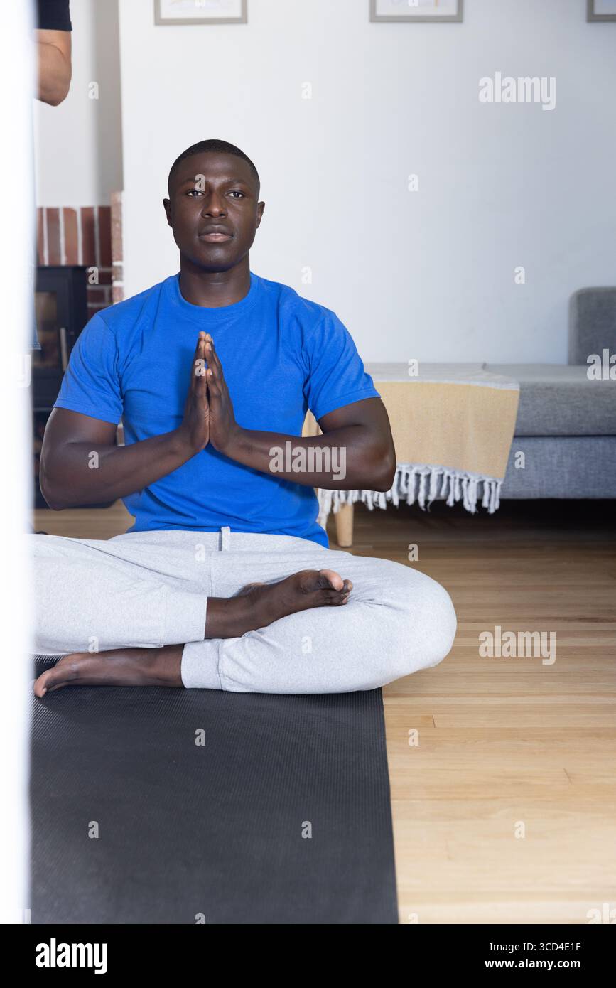Uomo afroamericano seduto e meditando sul tappetino nero da yoga nel soggiorno con t-shirt blu Foto Stock