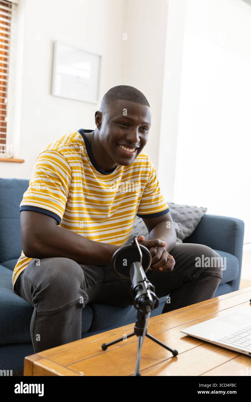 Uomo afroamericano seduto su un divano blu nel soggiorno utilizzando il microfono sul treppiede con un computer portatile Foto Stock