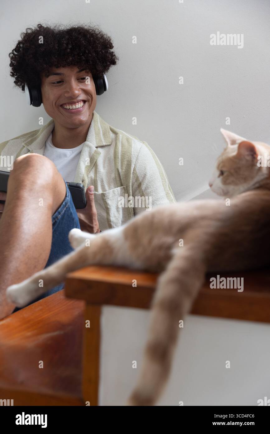 Uomo che indossa le cuffie con un tablet seduto in salotto accanto al gatto su un tavolo di legno Foto Stock