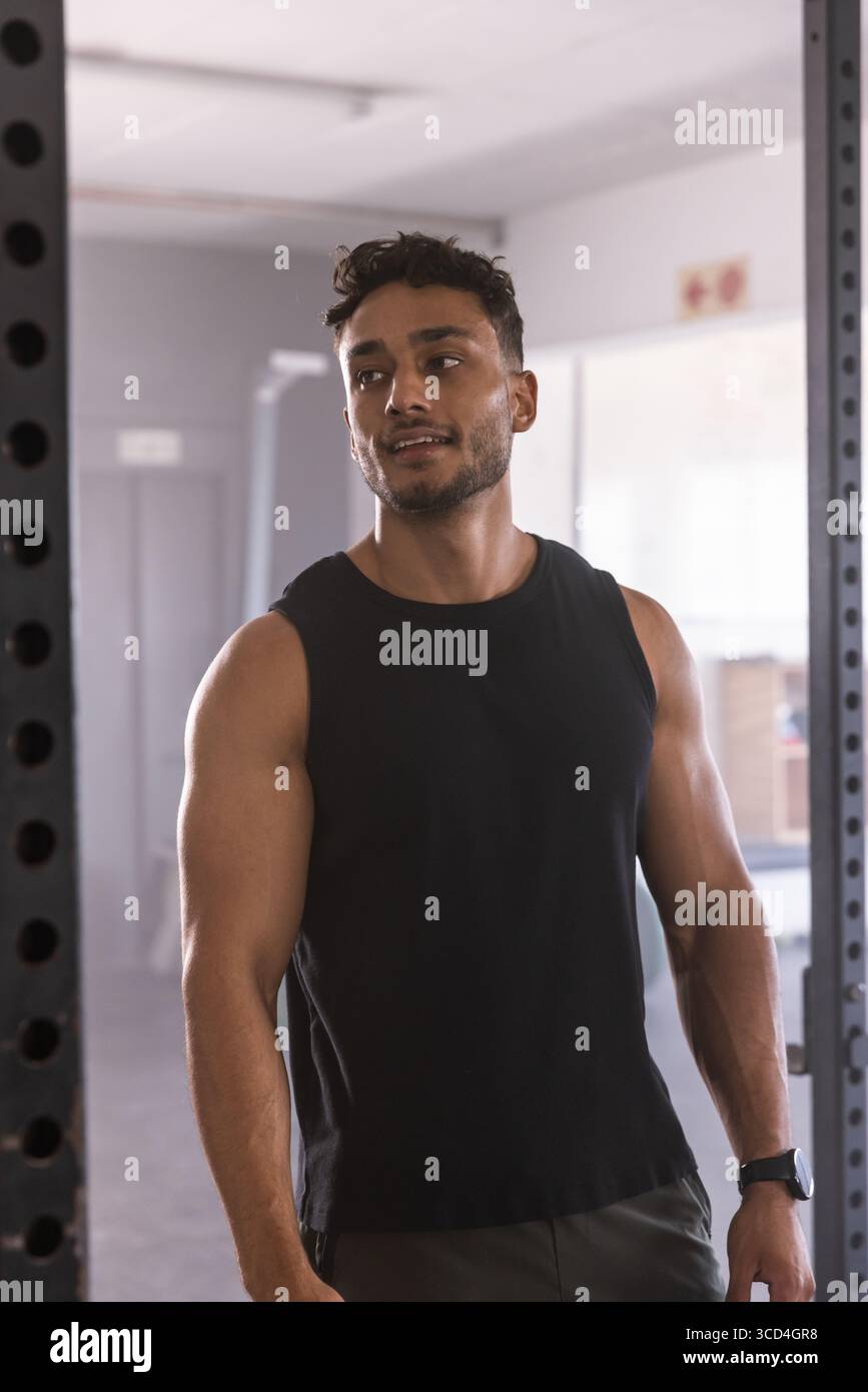 Uomo muscoloso in piedi accanto allo squat rack in palestra cattura il respiro indossando canotta e smartwatch Foto Stock