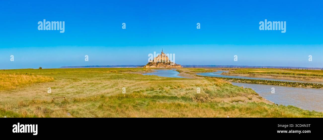 Un ampio panorama di dolci pascoli si estende sotto un cielo azzurro e cristallino, che conduce l'occhio al famoso isolotto roccioso della Normandia, Mont-Saint-Michel, a... Foto Stock