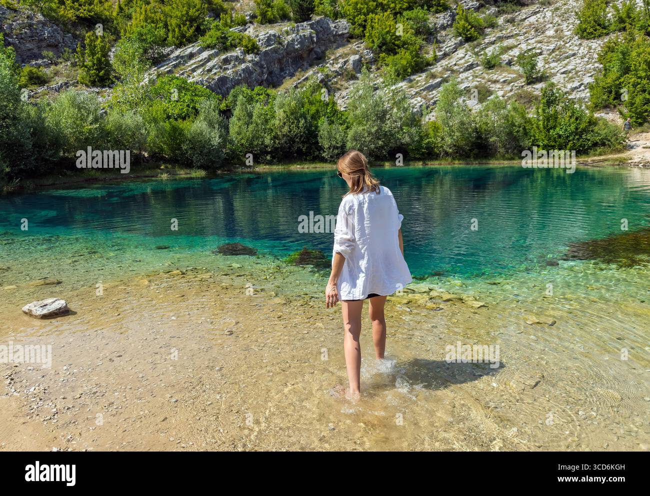 Una donna con una camicia bianca gode di una natura incontaminata e si immerge a piedi nudi nel lago di montagna cristallino Eye of the Earth of the Cetina River Foto Stock
