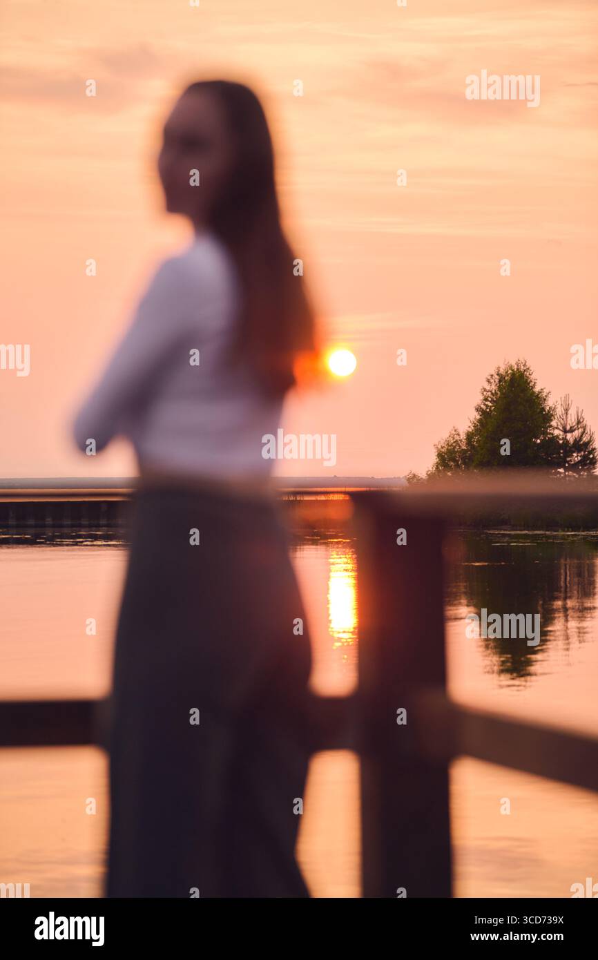 La silhouette femminile sfocata si affaccia sul tranquillo tramonto sul lago. Sfumature di rosa e arancione creano un'atmosfera tranquilla. L'albero e l'orizzonte distante aggiungono profondità a S. Foto Stock