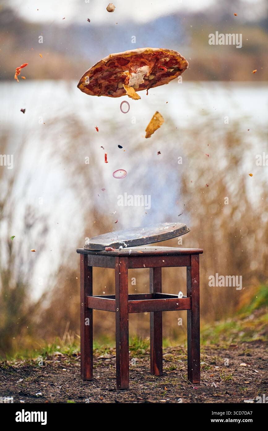Una vivace esplosione di pizza catturata a mezz'aria sopra uno sgabello rustico in legno all'aperto. I condimenti e i frammenti di crosta si diffondono drammaticamente contro il lago An sfocato Foto Stock Una vivace esplosione di pizza catturata a mezz'aria sopra uno sgabello rustico in legno all'aperto. I condimenti e i frammenti di crosta si diffondono drammaticamente contro il lago An sfocato Foto Stock