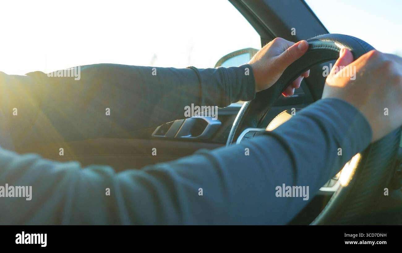 Le mani tengono saldamente il volante di un veicolo, illuminato dalla luce solare intensa, sottolineando la concentrazione e il viaggio in avanti in un ambiente dinamico Foto Stock