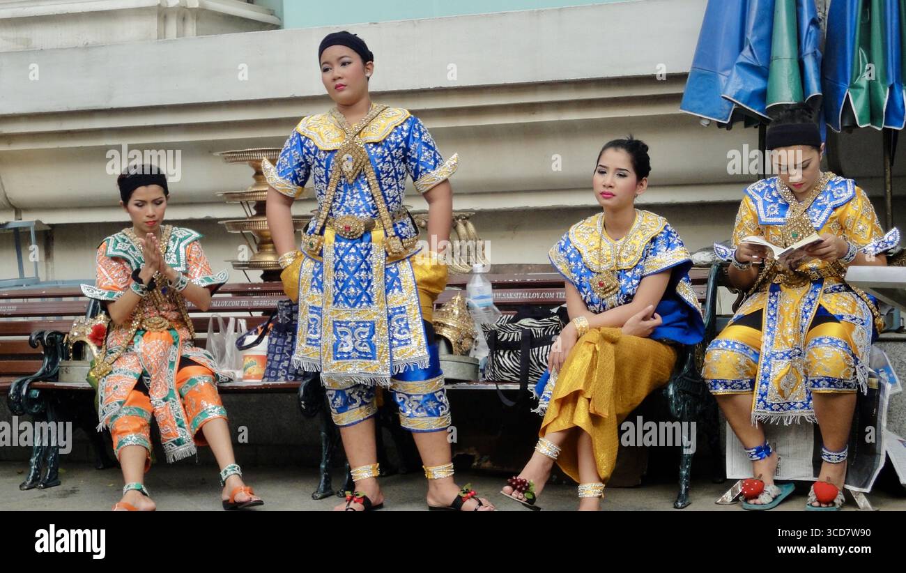 Ballerini thailandesi in costumi tradizionali decorati che riposano su una panchina a Bangkok, mostrando momenti di puro divertimento dietro gli spettacoli culturali. Foto Stock