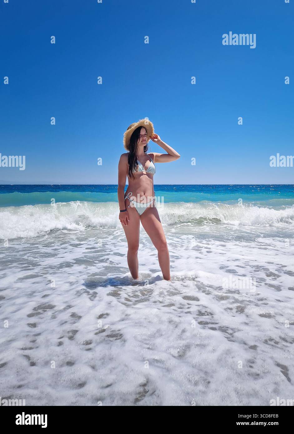Bella donna che posa in bikini e cappello di paglia, si erge tra le onde spumeggianti del mare azzurro, sotto un cielo estivo senza nuvole. Vacanze estive Foto Stock