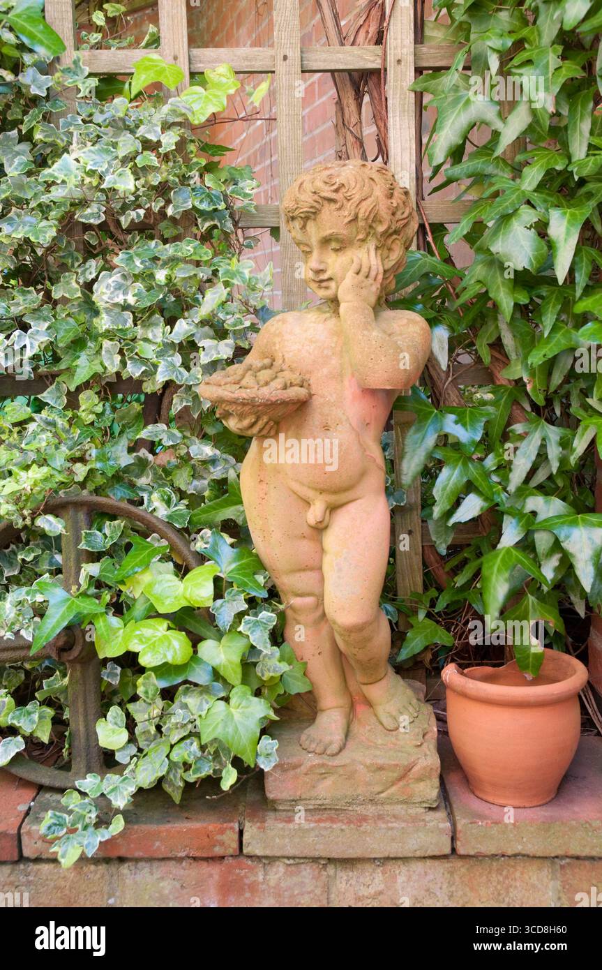 Una giocosa statua di un bambino si erge con cura accanto al vibrante edera verde e alle piante in vaso in un tranquillo giardino, catturando un momento sereno. Foto Stock