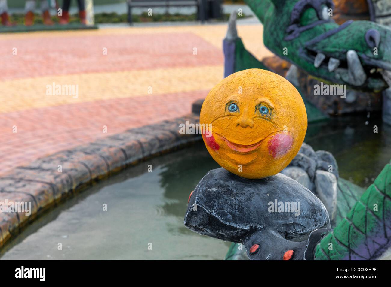 La stravagante scultura gialla del volto sorridente si trova sul bordo della fontana, presenta colori vivaci e un'atmosfera giocosa, perfetta per progetti creativi allegri Foto Stock