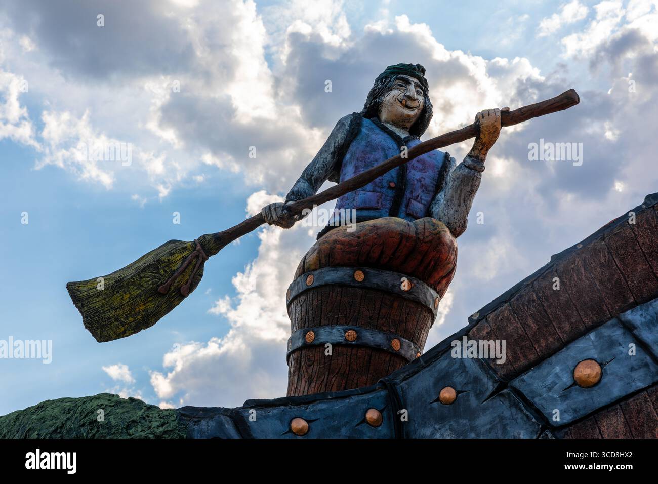 Statua colorata di una strega che regge una scopa, seduta su una canna, posta contro un cielo nuvoloso. Perfetto per temi folcloristici, Halloween o fantasia. Foto Stock