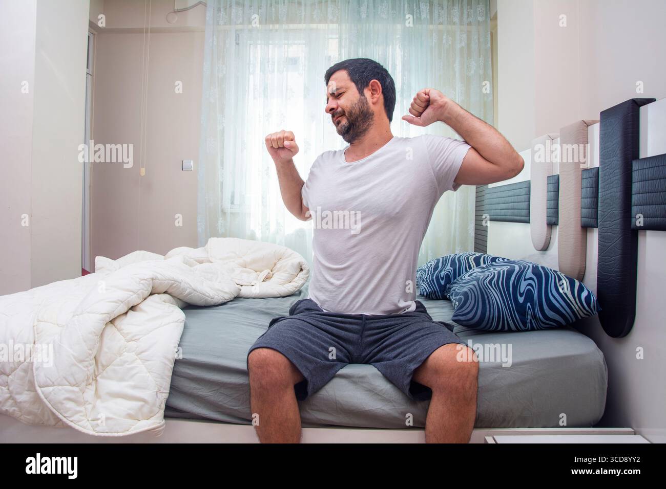 Un uomo che si è appena svegliato allunga il corpo seduto sul letto Foto Stock