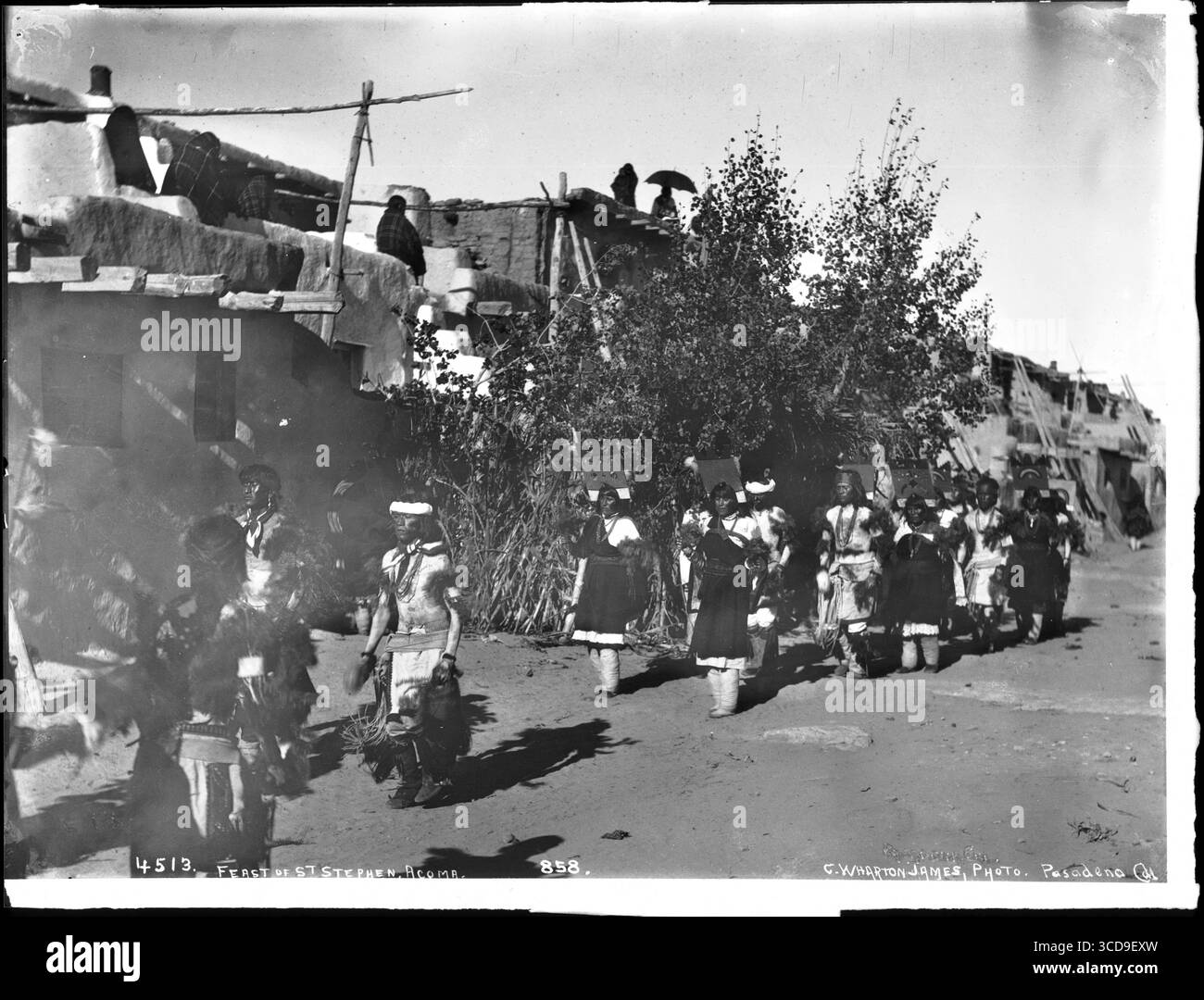 Fiesta de San Esteban (Santo Stefano), Acoma Pueblo, CA. 1900. I circa una dozzina di nativi americani Acoma indossano indumenti tradizionali, tra cui elaborati pezzi per la testa. Portano strumenti e piume in mano mentre ballano. L'edificio dietro i ballerini è realizzato in adobe con travi di coda in legno a vista. Le scale si appoggiano a intervalli regolari contro il lungo edificio. Un grande albero cresce al centro. Alcune persone osservano dal tetto. I ballerini ballano in una processione lungo la strada accanto all'edificio. Foto Stock