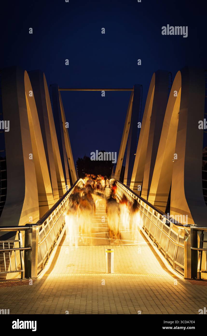 Vista notturna del ponte pedonale Wave of Azov a Kiev, Ucraina, con sagome sfocate di persone in movimento sotto luci calde contro un blu profondo Foto Stock