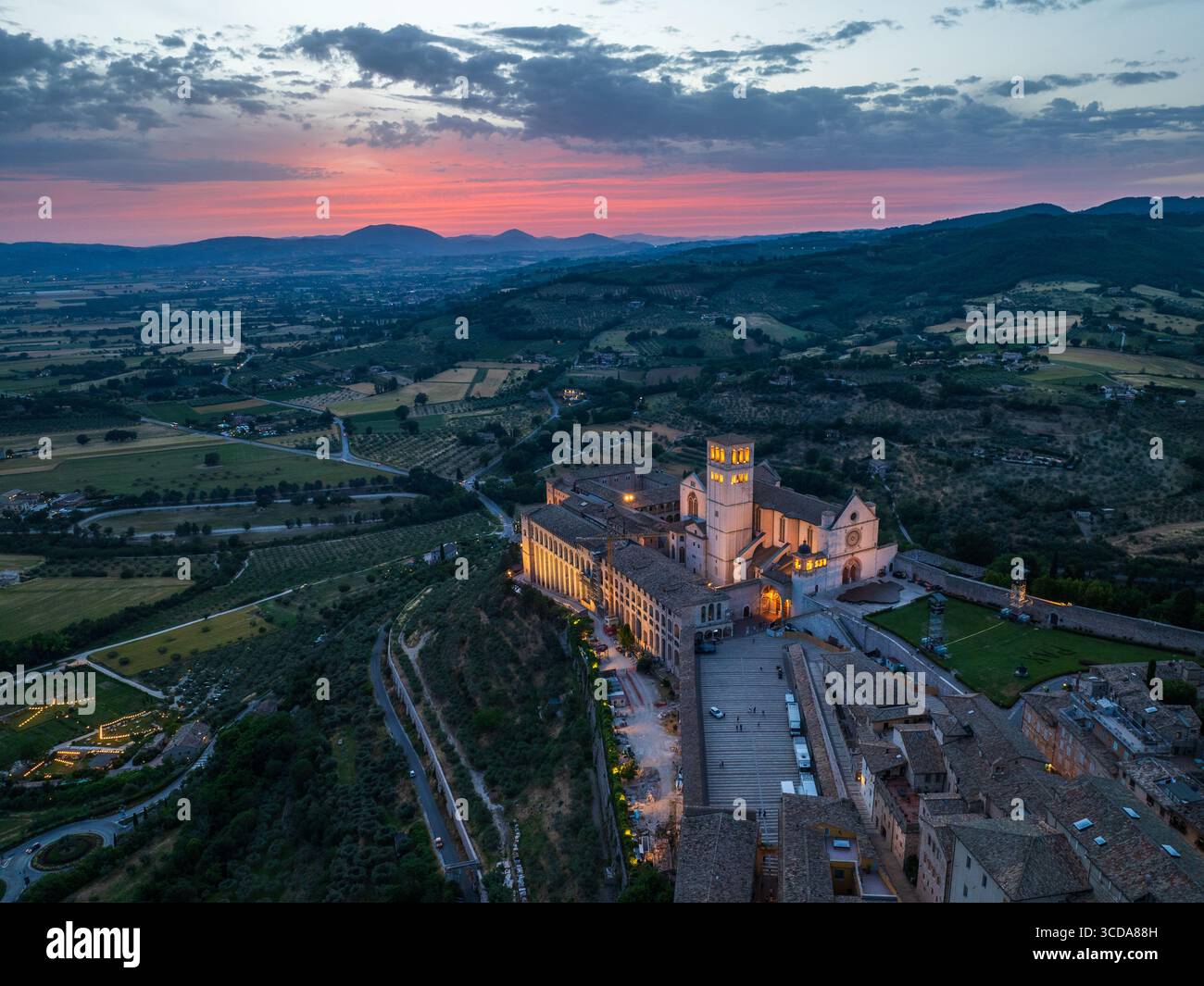 La vista aerea della Basilica di San Francesco d'Assisi si illumina con calore contro il cielo crepuscolare, un faro tra le dolci colline di Perugia, Umbria, Italia. Foto Stock