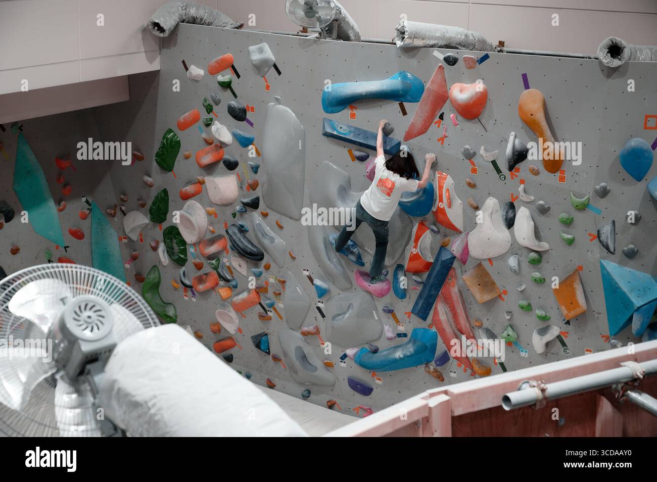 Arrampicatore che affronta una colorata parete di bouldering interna in una palestra dall'aspetto industriale con condotti esposti, ventilatori e ringhiere in metallo Foto Stock