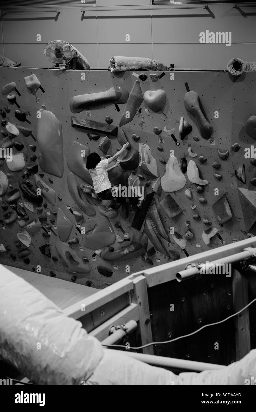 Vista monocromatica di una palestra di bouldering interna con un aspetto industriale, mentre un arrampicatore scala grandi, testurizzate, si tiene sotto i condotti esposti e i ventilatori montati Foto Stock