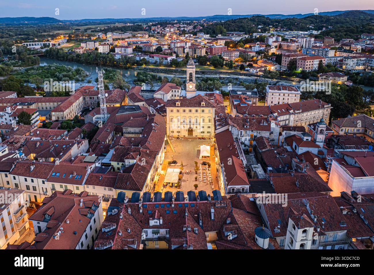 Vista aerea del Palazzo Civico che proietta un bagliore dorato su una vivace piazza tra i tetti in terracotta e il sinuoso fiume Dora Baltea, Ivrea, Piemonte, Italia. Foto Stock