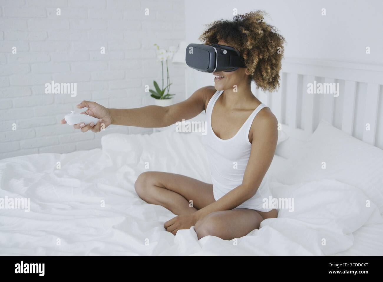Splendida modella giovane in costume da casa che si rilassa sul letto al mattino e indossa un visore VR eccitato da una nuova esperienza Foto Stock
