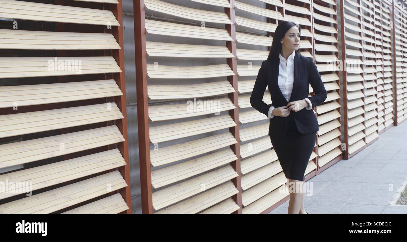 Elegante donna d'affari in fretta che cammina accanto a un edificio commerciale che abbellisce la giacca e guarda di lato Foto Stock