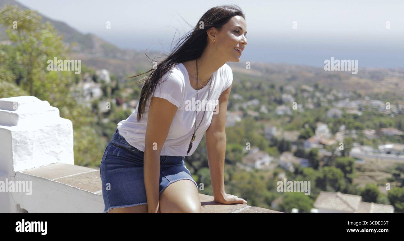 Bella bruna in un abito casual che sembra eccitato mentre posa sul punto panoramico ed esplora la città costiera dall'alto in estate Foto Stock