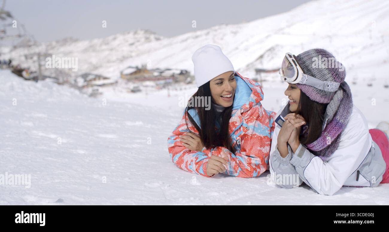 Graziose coppie di gemelle con lunghi capelli neri e abbigliamento da sci che si divertono a sdraiarsi sul fondo della pista da sci con la montagna alle loro spalle Foto Stock