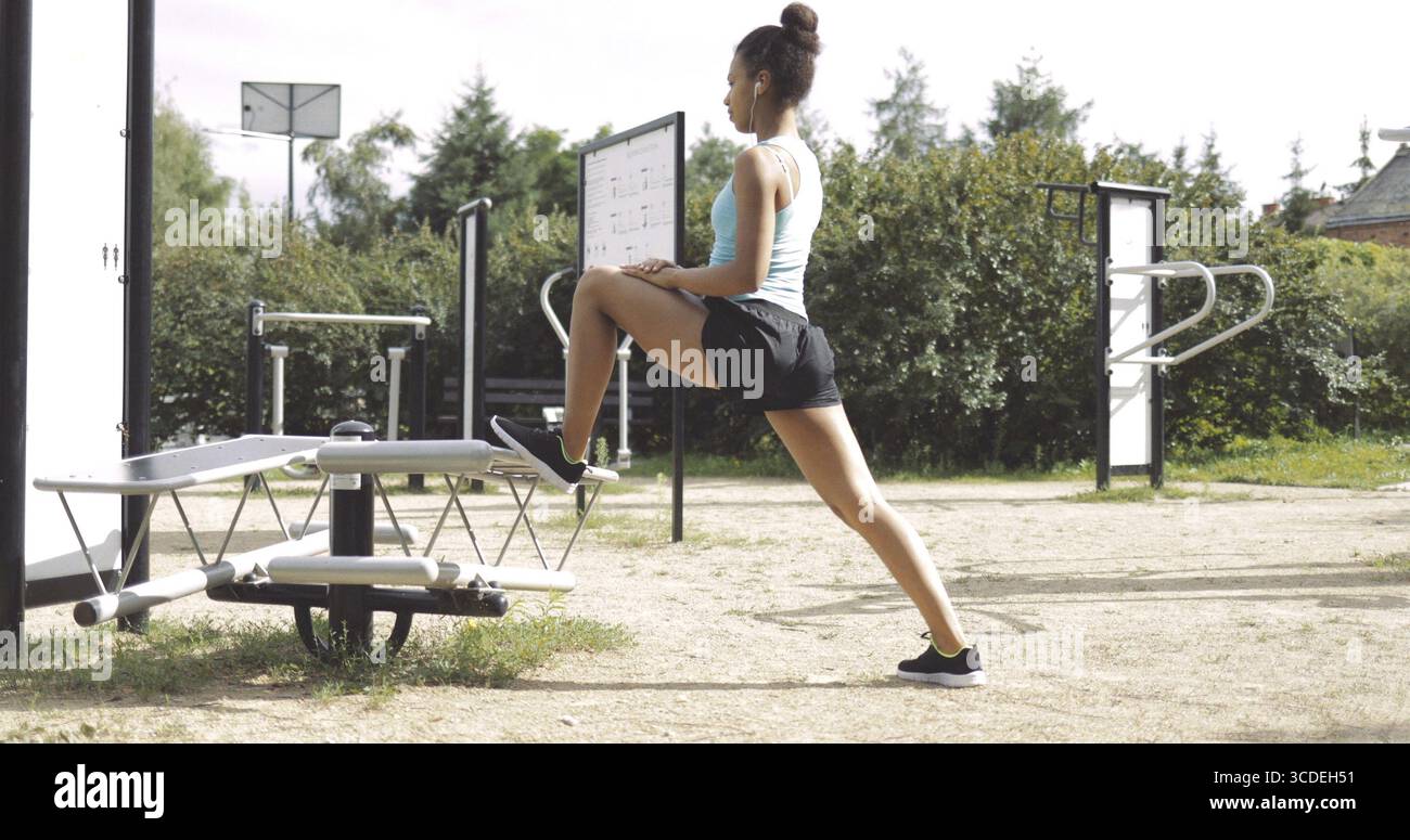 Vista laterale della donna in abbigliamento sportivo che ascolta la musica e si allunga le gambe mentre si allena nella palestra in strada nel parco guardando avanti Foto Stock