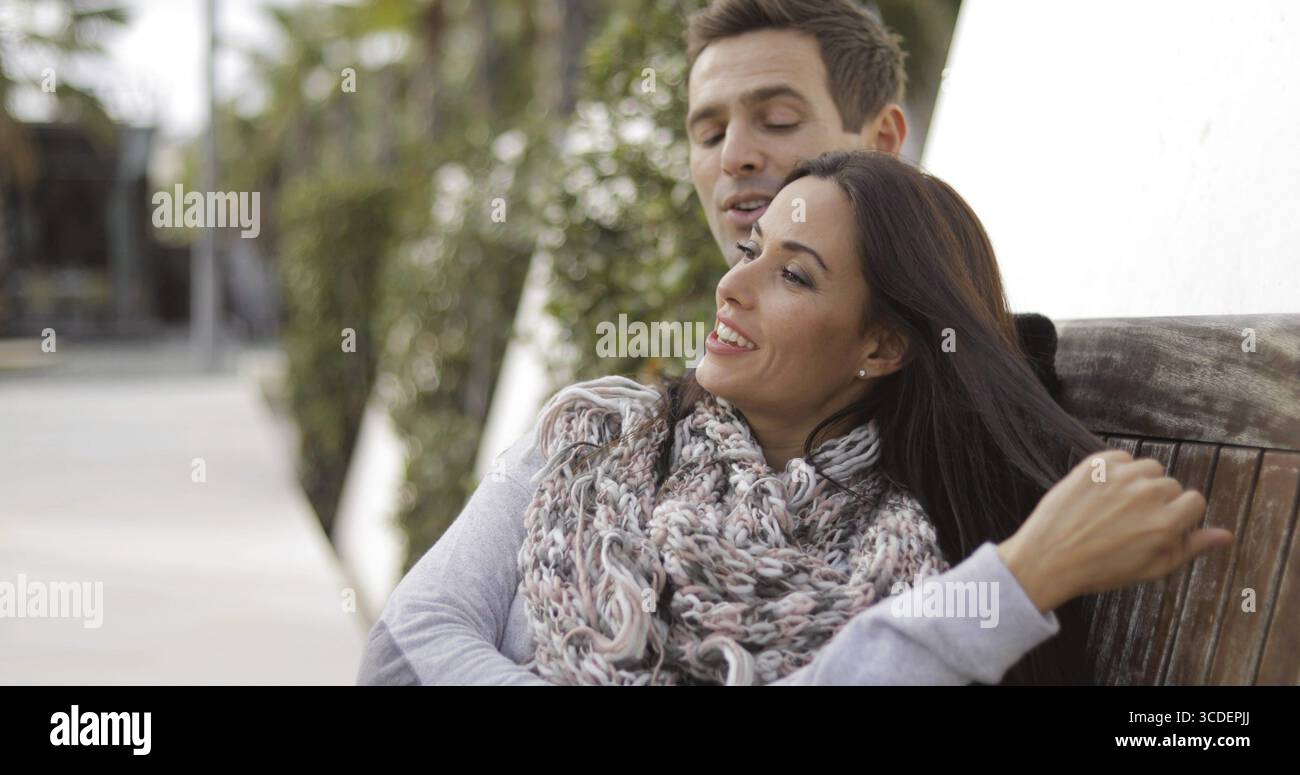 Giovane donna attraente e alla moda che si rilassa con il suo beau mentre si siede insieme su una panchina di legno del parco guardando in lontananza con un sorriso focalizzato su di lei Foto Stock