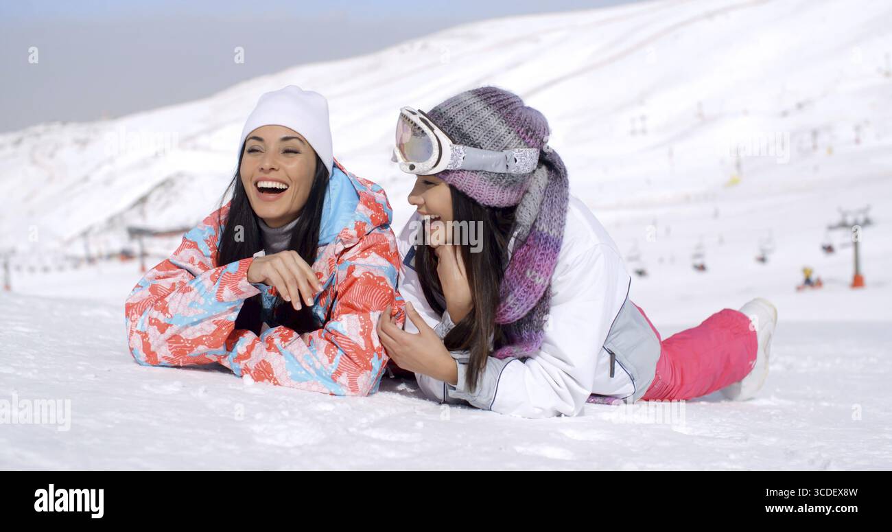 Simpatici gemelli con abbigliamento da sci che ridono mentre si stendono a terra in fondo alla pista con la montagna alle loro spalle Foto Stock