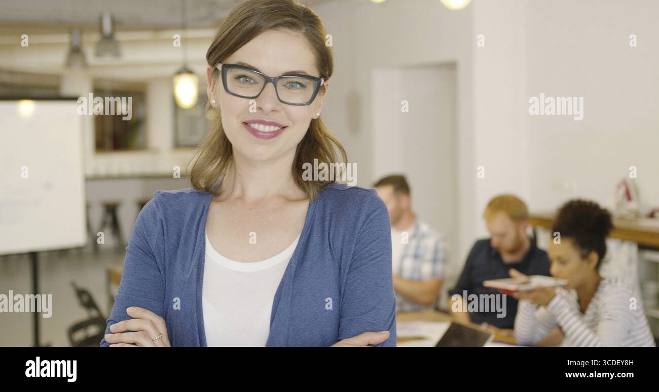 Ritratto di una giovane bruna che indossa abiti formali e occhiali da vista neri che posa con le mani incrociate e sorride alla macchina fotografica in ufficio Foto Stock