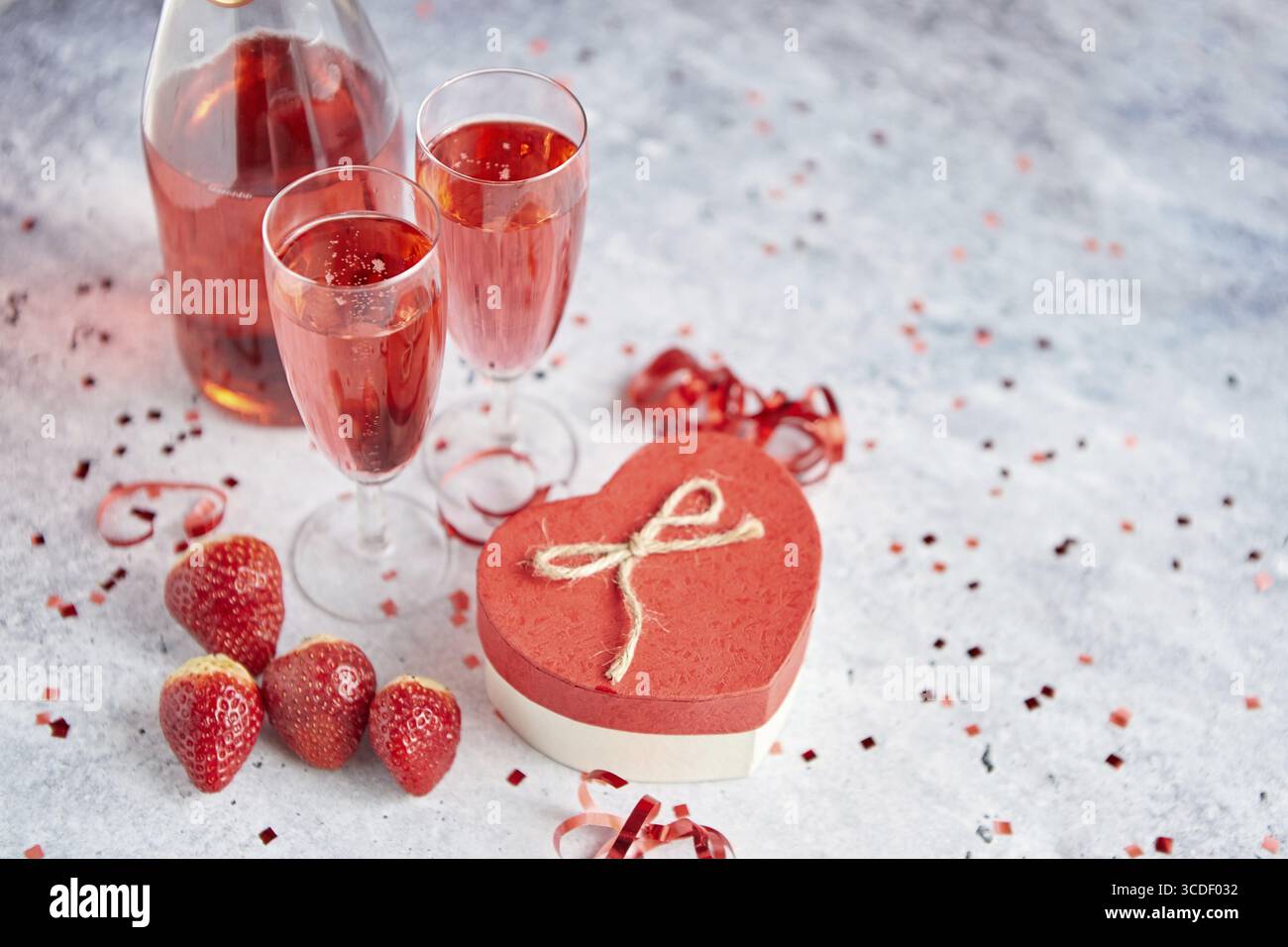 Bottiglia di champagne rosa, due bicchieri con fragole mature fresche e omaggio a forma di cuore, collocati su un tavolo in pietra per un'occasione speciale e romantica Foto Stock