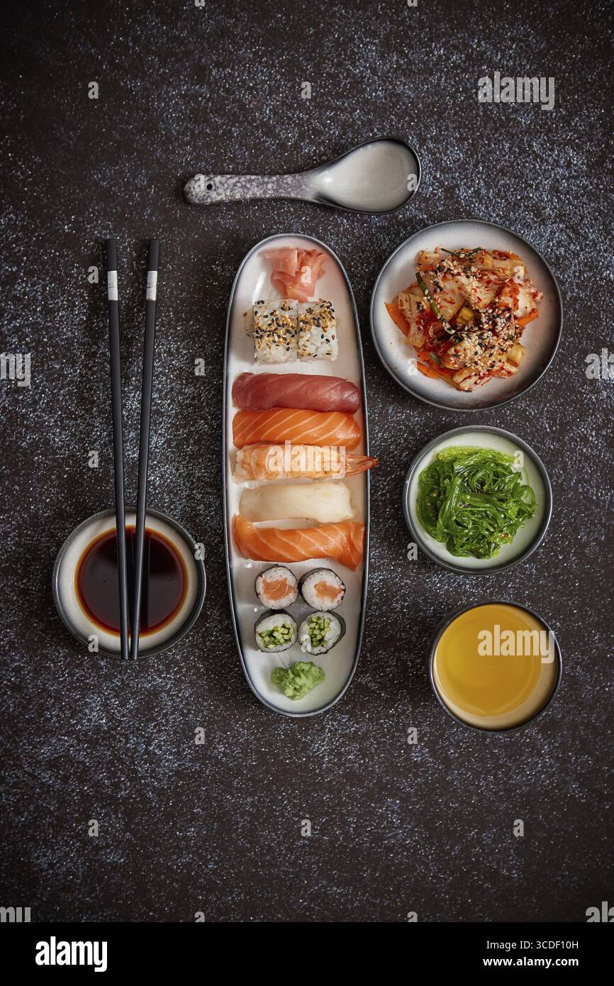 Assortimento di piatti asiatici. Vari sushi roll posizionati su piatti di ceramica. Insalate kimchi e goma wakame. Zuppa di soia e bacchette ai lati. bac scuro e grungy Foto Stock